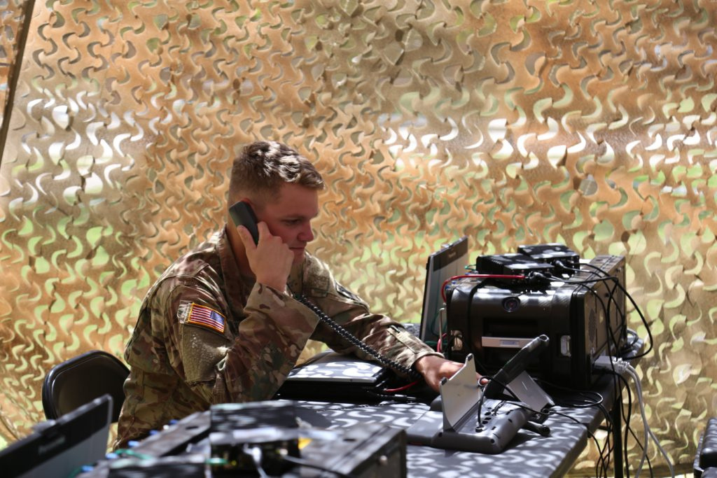 A focused military personnel working on communication solutions while making a phone call in a tent with a camouflaged background.